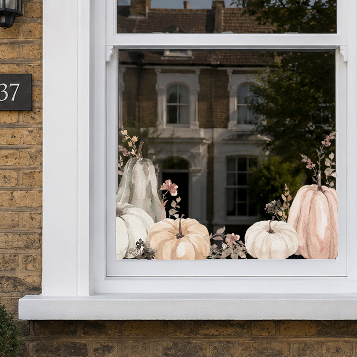 A set of watercolor-style fall window decals featuring pumpkins, gourds, and small flowers, shown applied to a white-framed window on a brick house exterior.